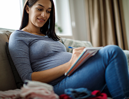 A pregnant woman sits on a couch and writes in a notebook.