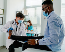 A doctor examines a patient's leg. Both are wearing masks.