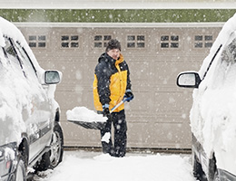 Snow falls heavily. A man in a yellow and black coat stands between two parked cars holding a big shovel full of snow. 