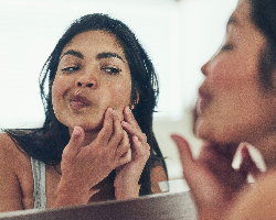 A dark-haired woman leans into her bathroom mirror, examining a spot on her cheek with both hands.