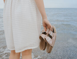 Photo of a woman standing on the beach, holding sandals.