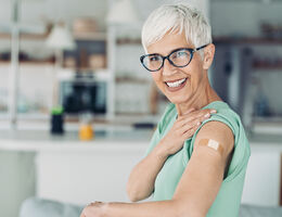 An older woman smiles and holds up her sleeve to show an adhesive bandage on her upper arm.
